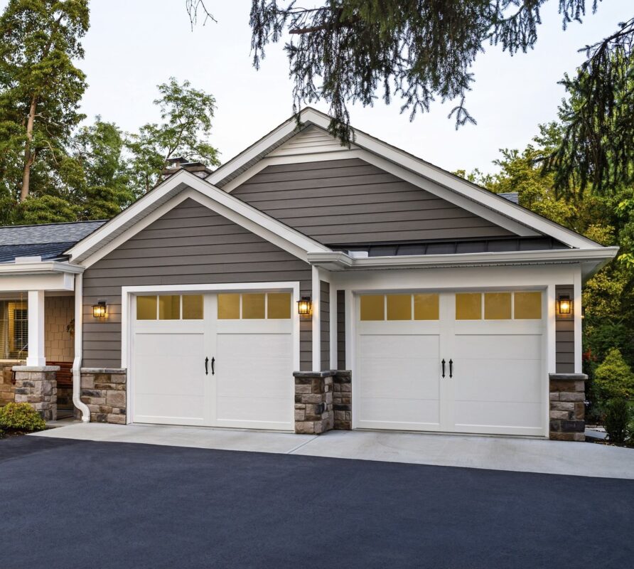 White garage doors with decorative windows and stone accents, enhancing home security features against remote hacks, surrounded by lush greenery.
